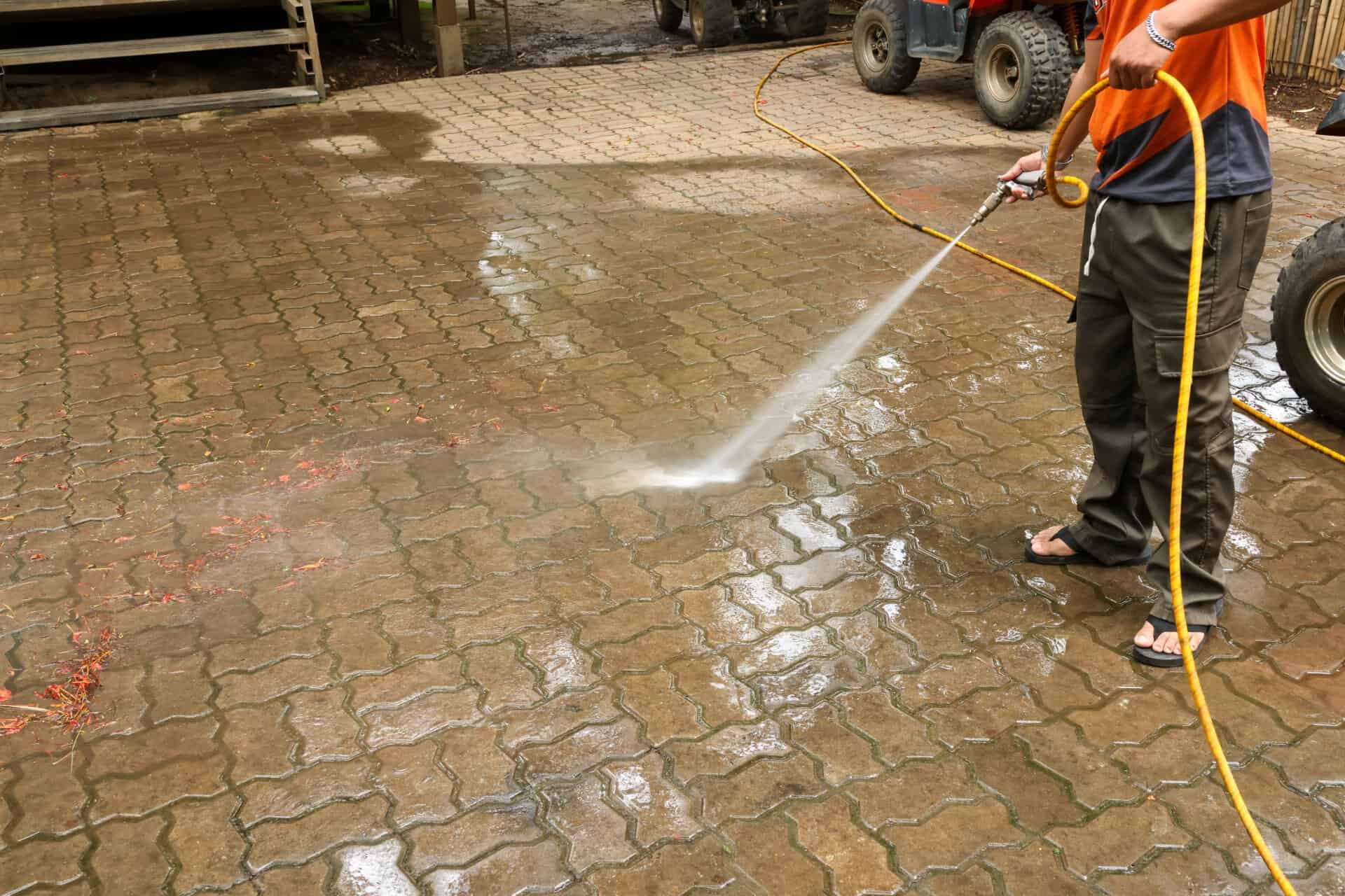 Focused worker operating high pressure washer equipment, cleaning dirty paving stone on residential driveway. Powerful water spray from yellow hose removes grime effectively outside